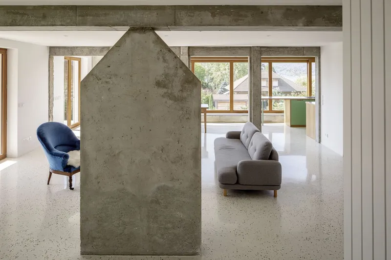 Living room from fireplace: house-shaped concrete chimney, grey sofa behind, timber doors, mountain light