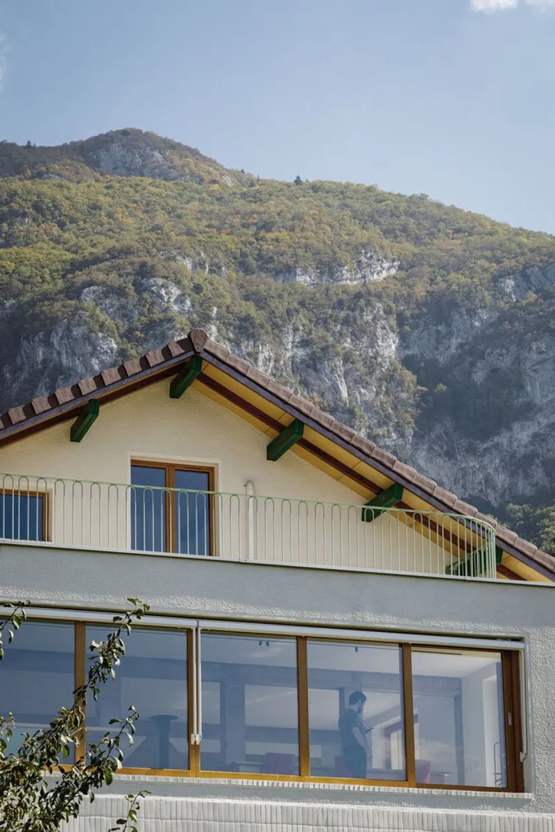 Upper floor detail: timber louvers in gable, yellow-painted rafter ends, mountain cliff behind