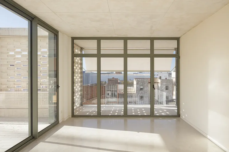 Living room interior: corner with floor-to-ceiling glazing, terrace beyond, exposed concrete ceiling, light grey floor, city view