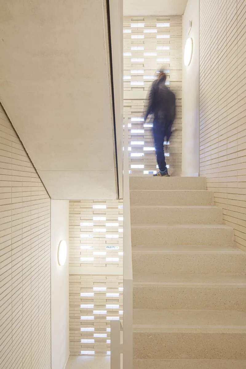 Stairwell: figure ascending concrete stairs, perforated ceramic screen casting patterned light on the walls, timber handrail