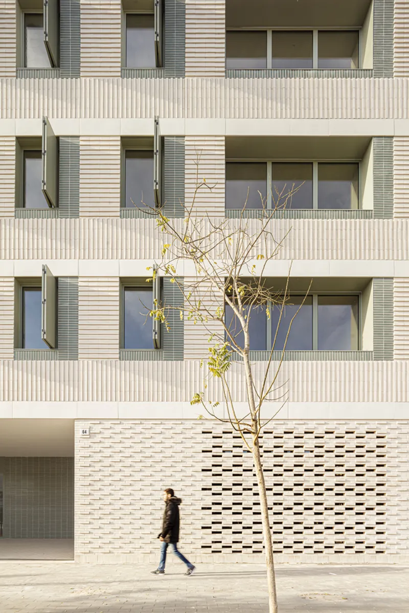 Street facade from the west: cream brick with perforated ceramic base, regular window grid, figure walking, young tree on the pavement