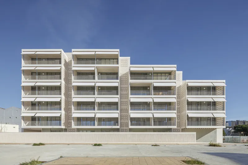 South-west facade: full-width view showing five levels of generous balconies, cream brick, stepped roofline, clear sky