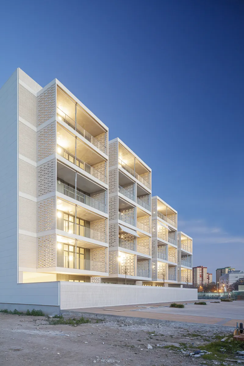 Corner at dusk: stepped upper levels catching the last light, deep balconies, cream brick, distant buildings at right
