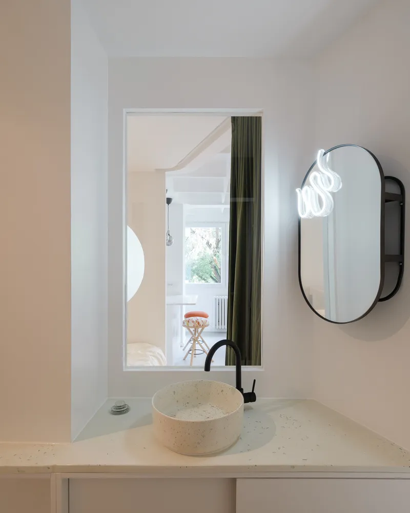 Bathroom: terrazzo basin on a white counter, rectangular and oval mirror composition, view back into the living room through the circular window reveal
