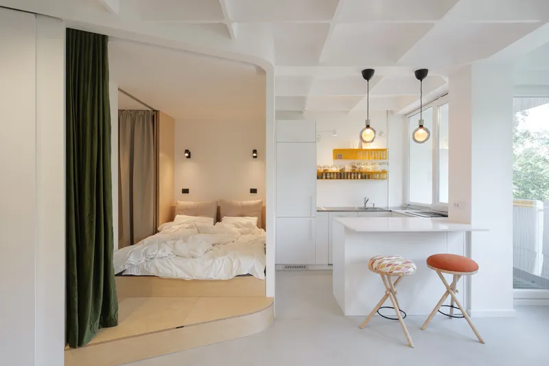 Wide view: kitchen with three pendant lights at right, bedroom platform raised above the main floor, green velvet curtain drawn back, white coffered ceiling