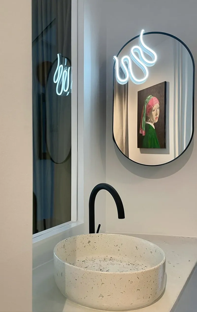 Bathroom accent: neon cursive sign reflected in the oval mirror, framed portrait in a pink frame, terrazzo basin below