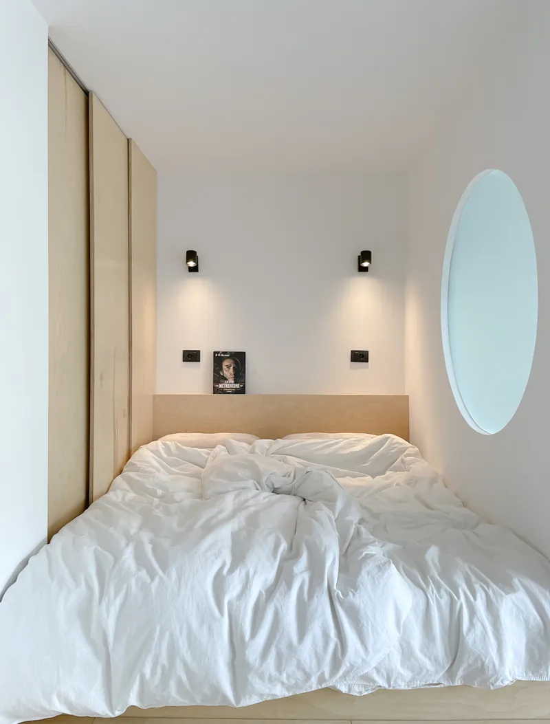 Bedroom close-up: white bedding and pillows, two matching black wall lamps, timber headboard, round window on the right wall