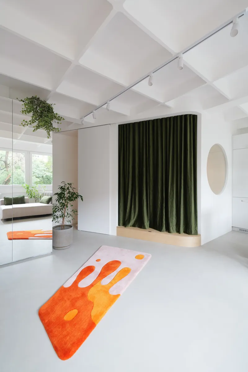 Living room with rug: orange-on-pink kite-shaped rug on the polished white floor, green velvet curtain behind, round pink mirror, plants cascading from above