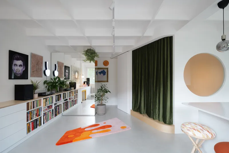 Main living area: white coffered ceiling, low built-in bookshelf along the wall, green velvet curtain at right, orange abstract rug, plants and mirror wall