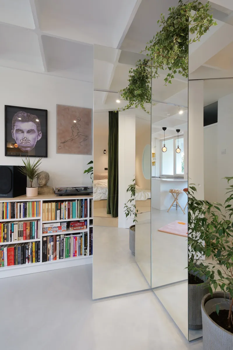 Mirrored wall: floor-to-ceiling mirror reflecting the bookshelf and living room, plants cascading from the ceiling, framed art on the left