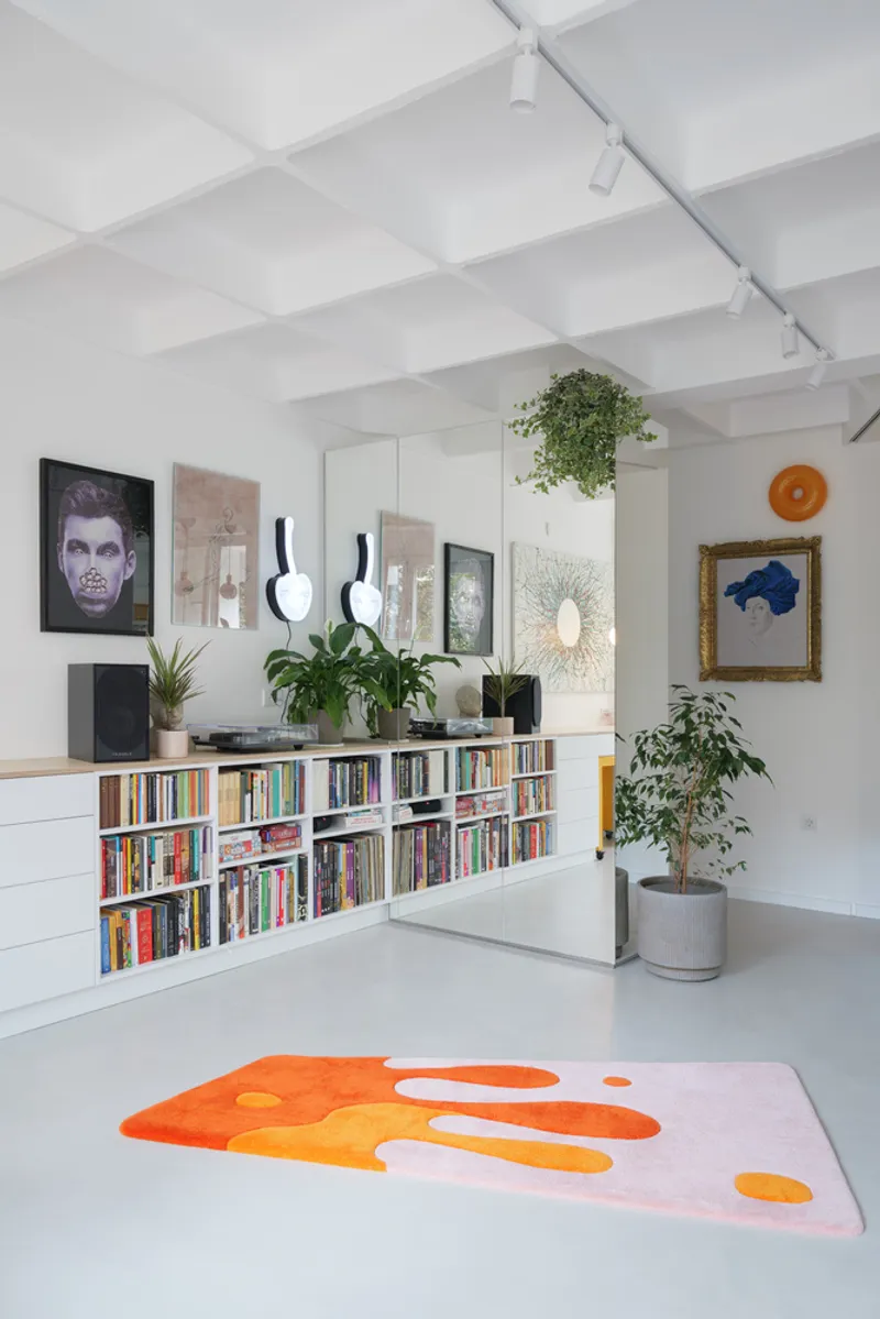 Living room wide view: long low bookshelf, white coffered ceiling, cascading plants, black artwork collection, orange rug on the white floor