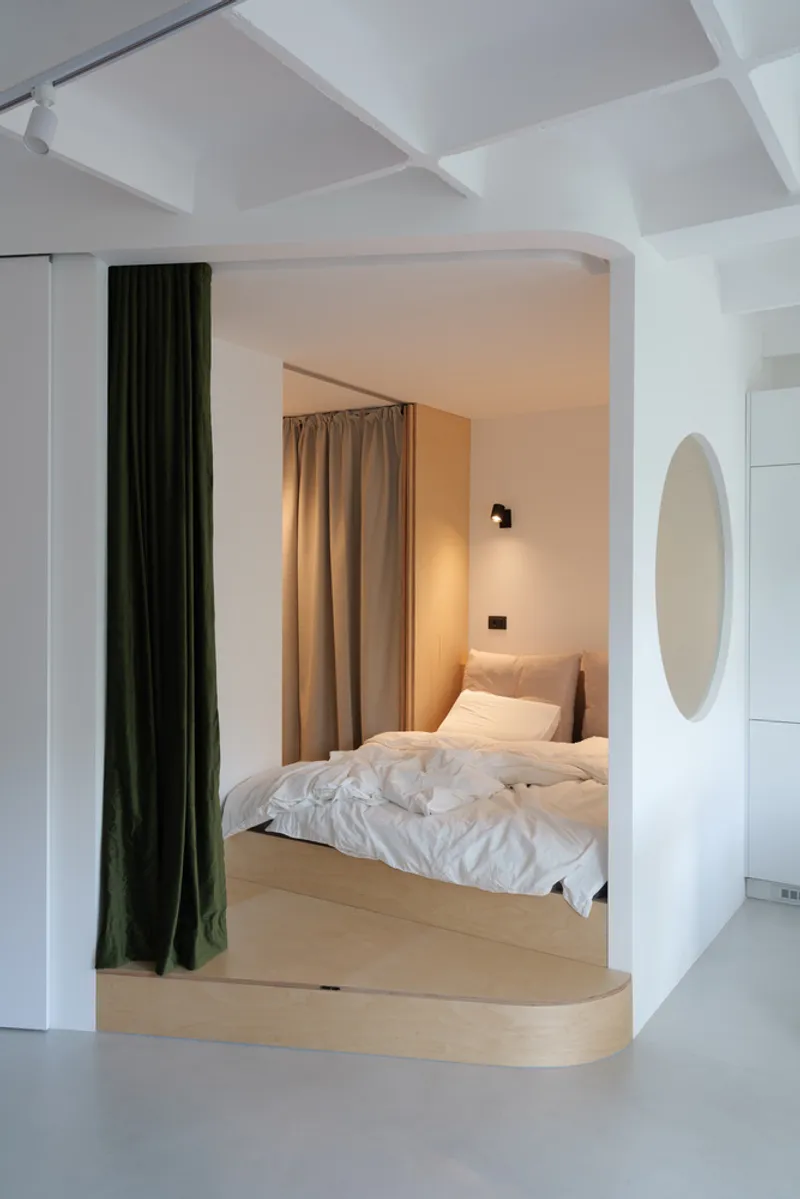 Embedded bedroom: raised timber platform with a white bed, green velvet curtain as a movable partition, round mirror on the wall, wall sconce