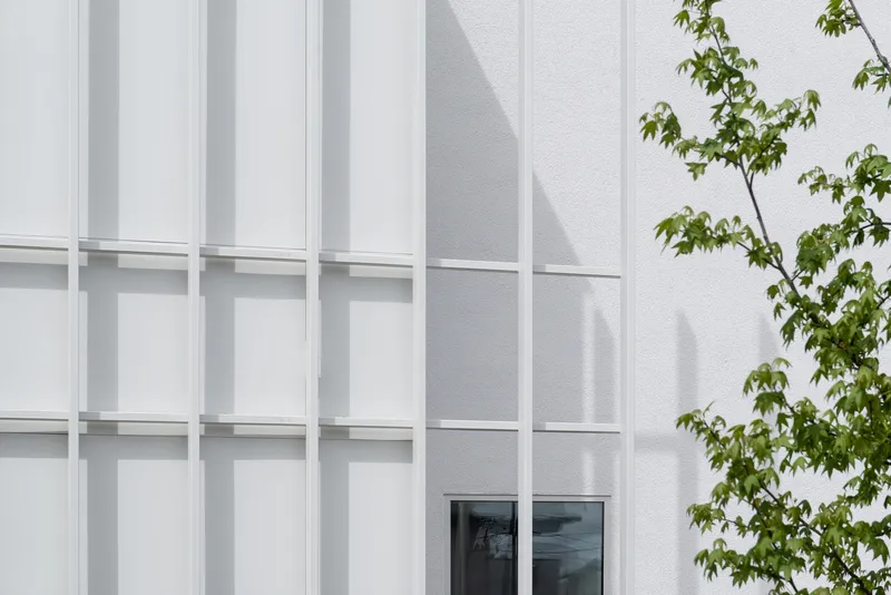 Facade close-up: white metal fin rhythm, small window set deep in the wall, tree shadow on the surface