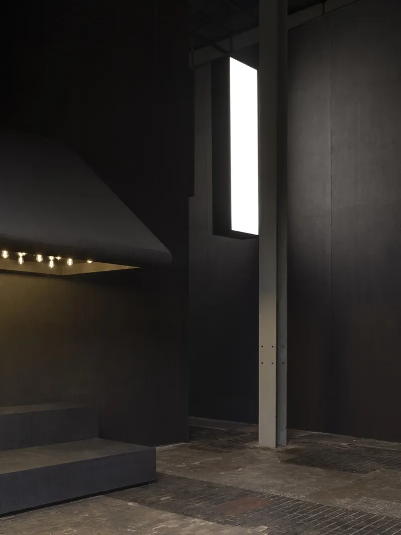 Vertical luminous slot window between dark wall panels in the gallery space