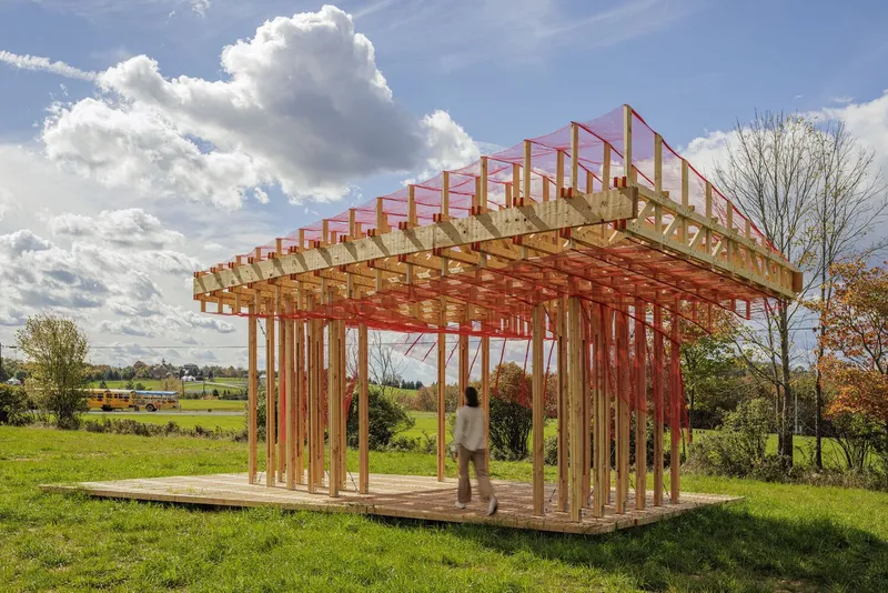 Main view: timber column and truss lattice with red construction-mesh curtains above, open timber deck, figure standing inside, cloudy sky over farmland