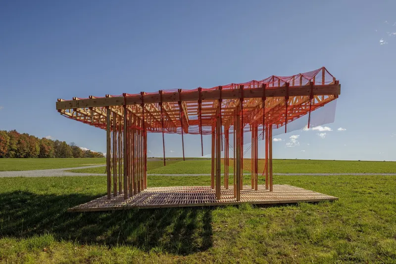 Side view: the full pavilion from the grass, timber truss roof, red painted stripes on the columns, autumn trees and sky beyond