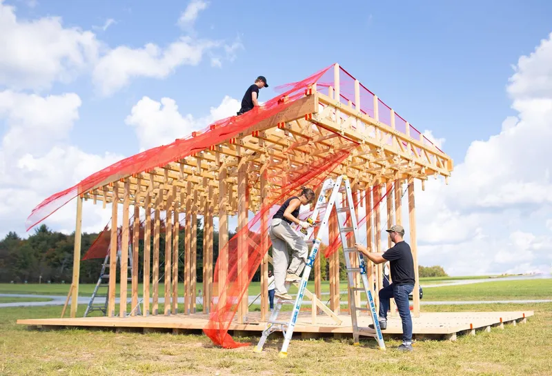 Construction: workers on ladders draping the red mesh curtain over the completed timber lattice, blue sky, open field