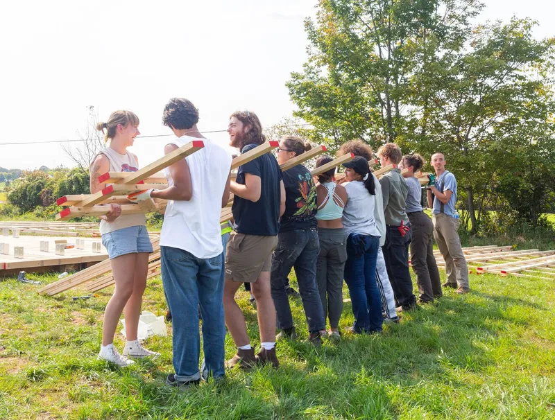 Assembly workshop: a line of students passing prefabricated timber truss members outdoors, prepped joints with red connectors visible in the foreground