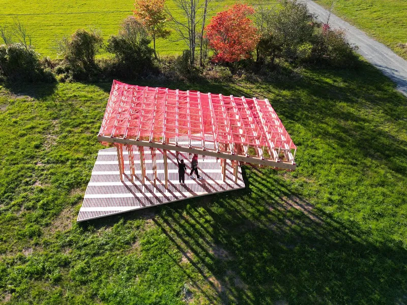 Aerial: the pavilion's red mesh roof from above with two figures inside, the truss pattern casting a striped shadow on the grass, autumn trees at the edge