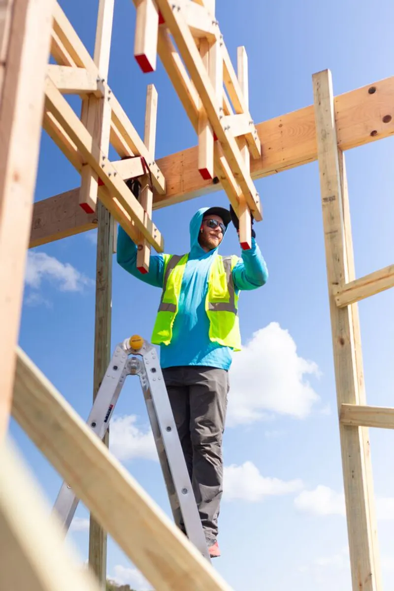 Assembly in progress: a worker in a hi-vis vest standing on a ladder, joining a timber truss bolt-by-bolt under a clear blue sky