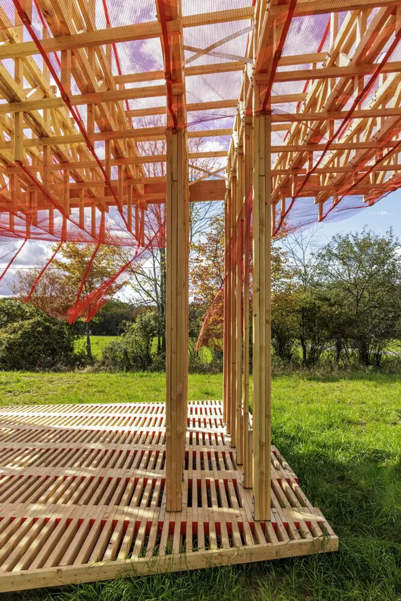 Interior: vertical timber slats forming a screen, timber lattice floor grating, figure in soft focus, meadow visible at the far end