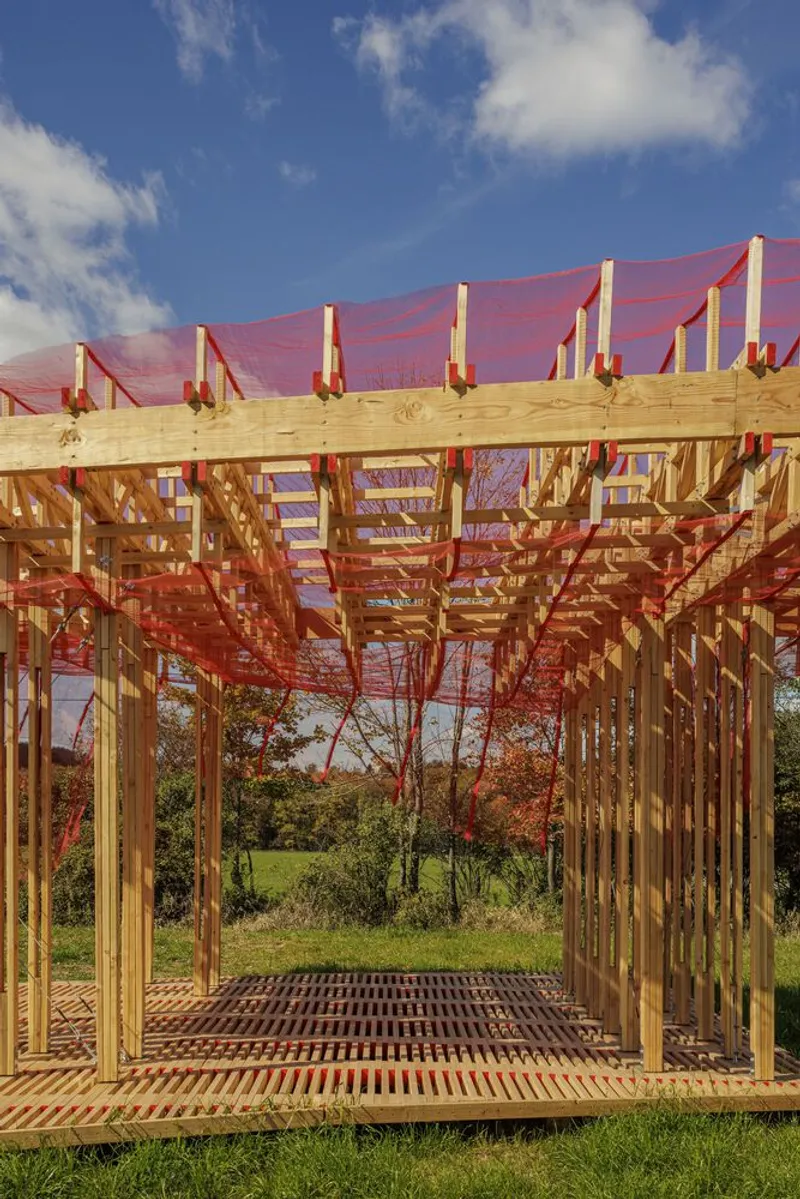 Three-quarter view: the full pavilion on its timber deck, red mesh above, autumn trees behind, warm evening light