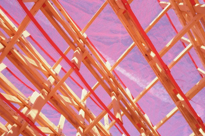 Interior roof: red mesh diffusing sunlight into a pink glow, cross-braced timber rafters, warm light on the timber structure