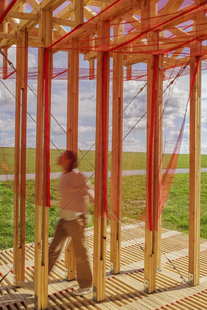 Interior with figure: person walking through the timber lattice, red mesh overhead, deck slats below, meadow and sky framed at the far end
