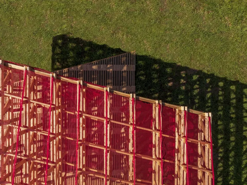Aerial corner: close-up of the red mesh roof over the timber lattice, black metal louvres at the edge, grass and shadow pattern visible below