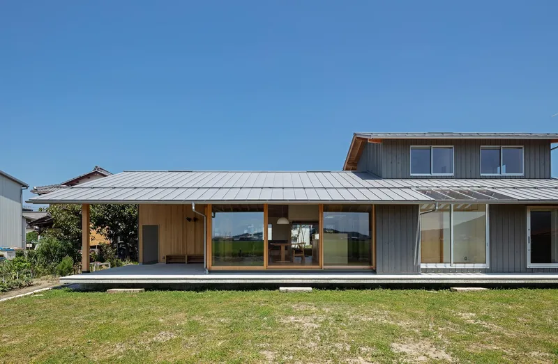 Garden side: single-storey cedar house with metal standing-seam roof, deep overhang, covered terrace, lawn, blue sky