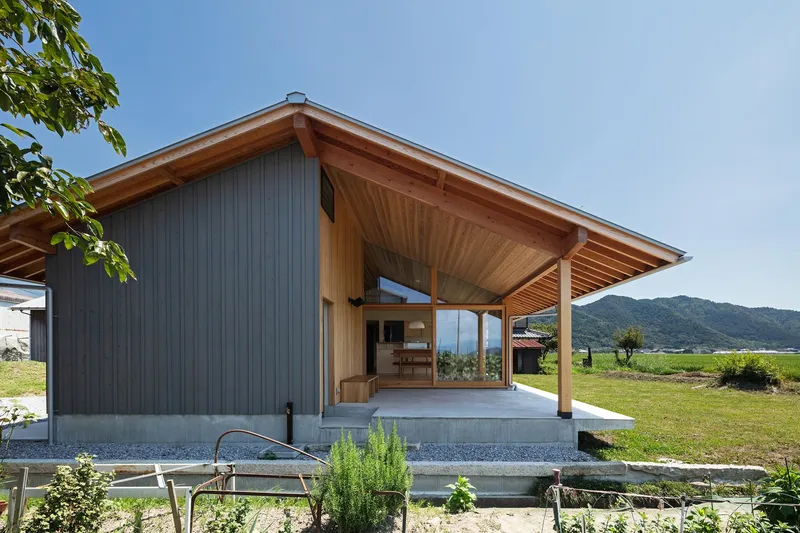 Entry side: pitched roof with deep cedar-lined overhang, metal cladding, concrete step, garden, mountains in background