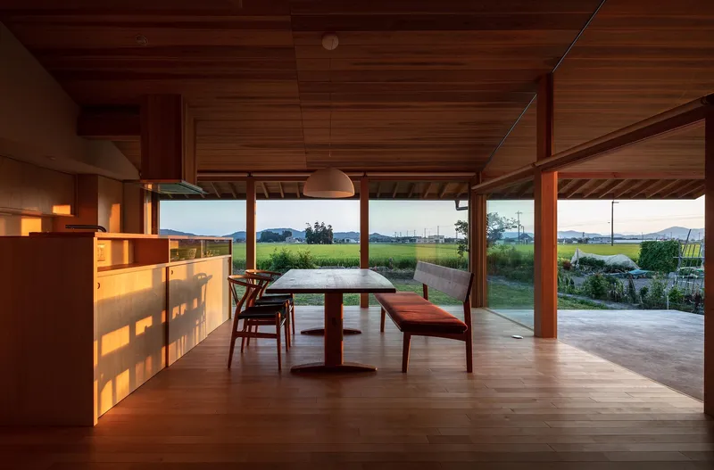 Dining at sunset: cedar table, leather bench, pendant lamp glowing, shelving lit warmly, rice fields darkening through the window