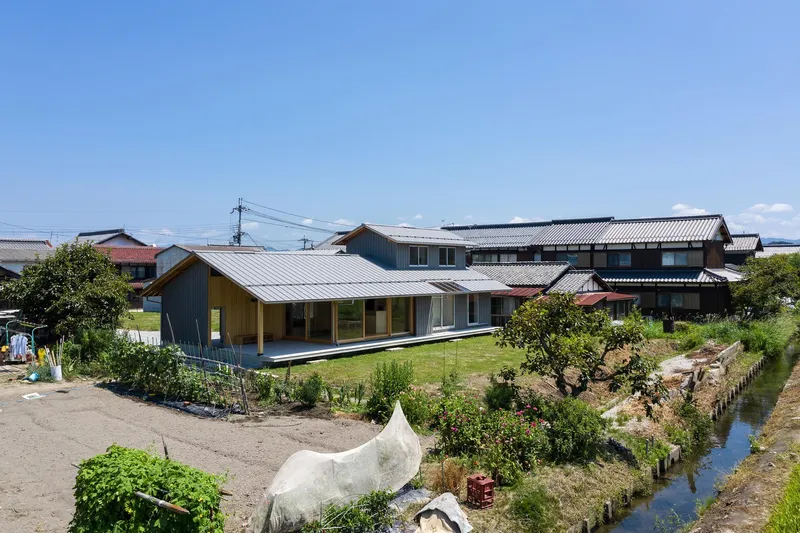 Village context: the house with its low metal roof sitting among traditional Japanese houses, stream, vegetable gardens