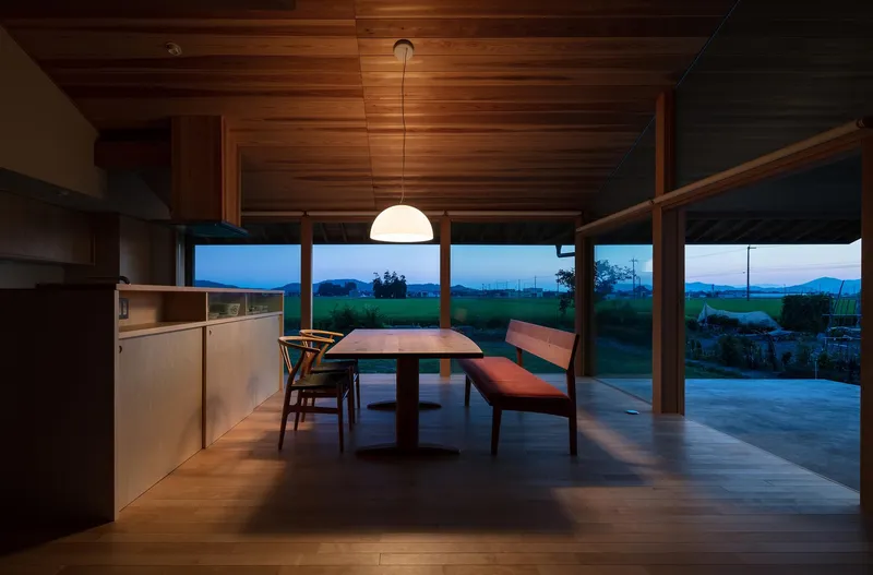 Dining at dusk: cedar interior glowing under pendant lamp, mountains silhouetted through full-width window, deep blue sky