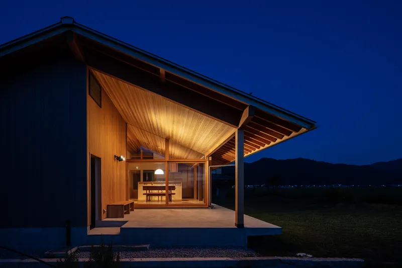 Night view from terrace side: lit cedar overhang, dining table and pendant visible through glass, mountains in twilight