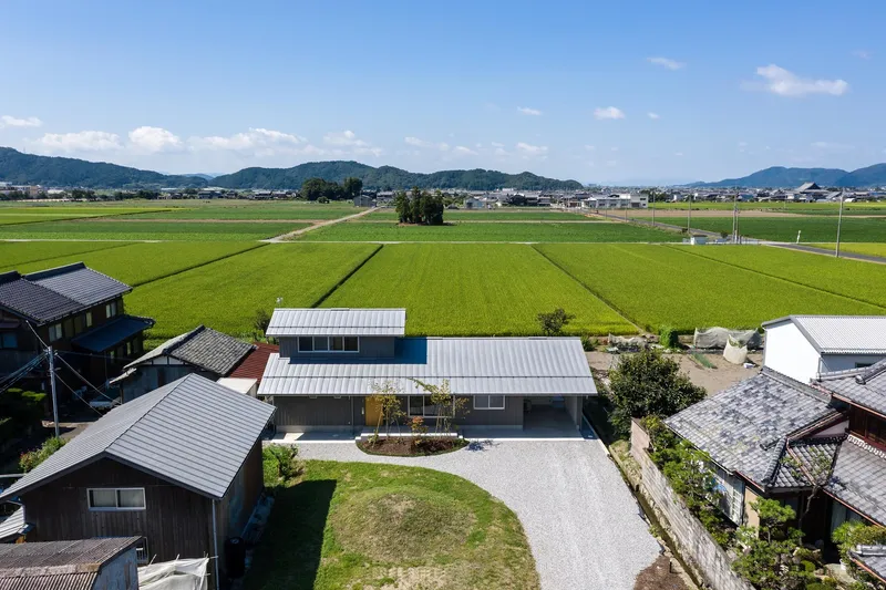 Aerial: the metal-roofed house set within the village, surrounded by bright green rice paddies stretching to mountains