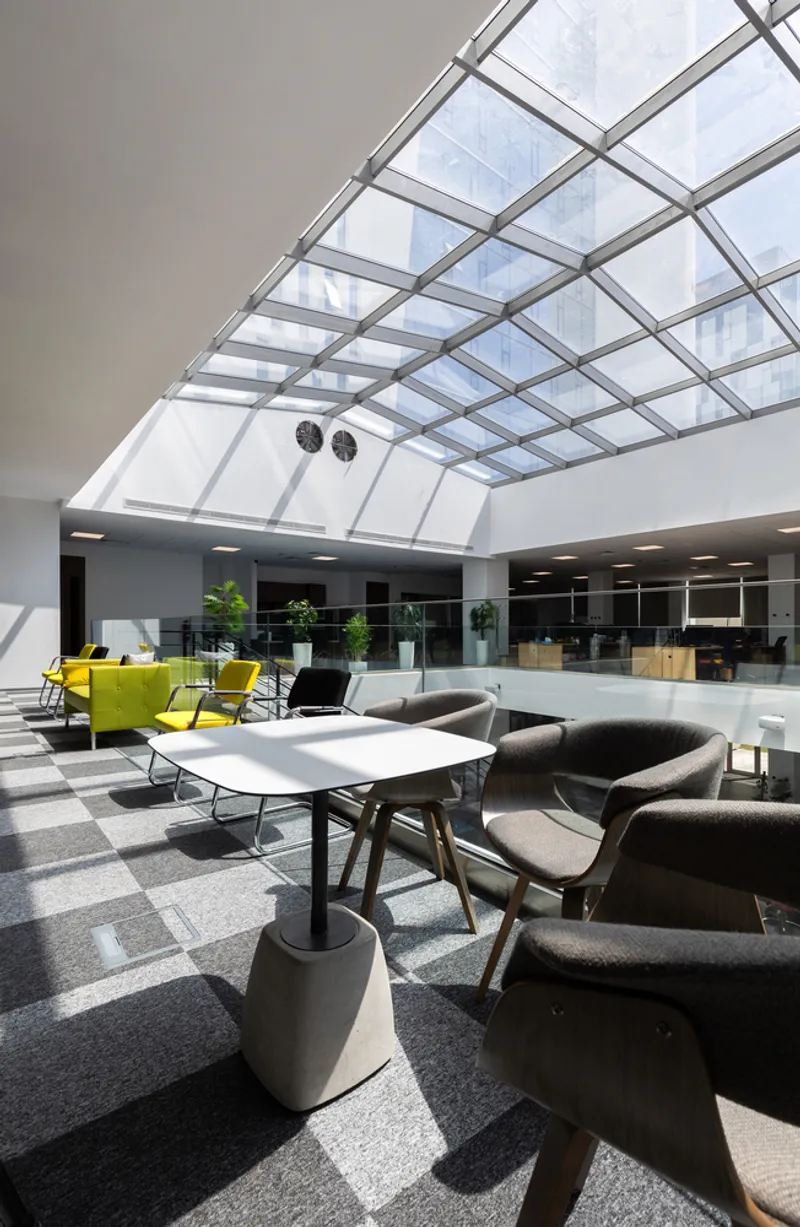 Atrium lounge beneath gridded skylight with seating clusters casting sharp shadows on terrazzo floor