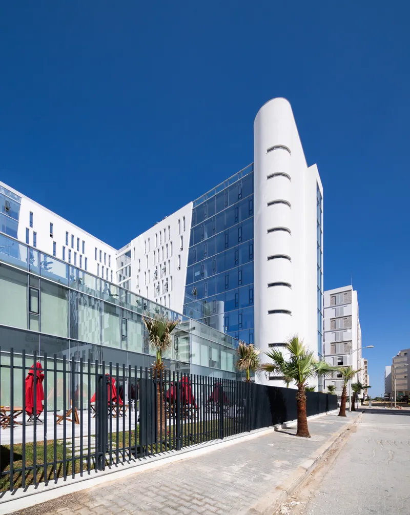 Street view of the white facade with punched windows and cylindrical glass tower under a clear blue sky