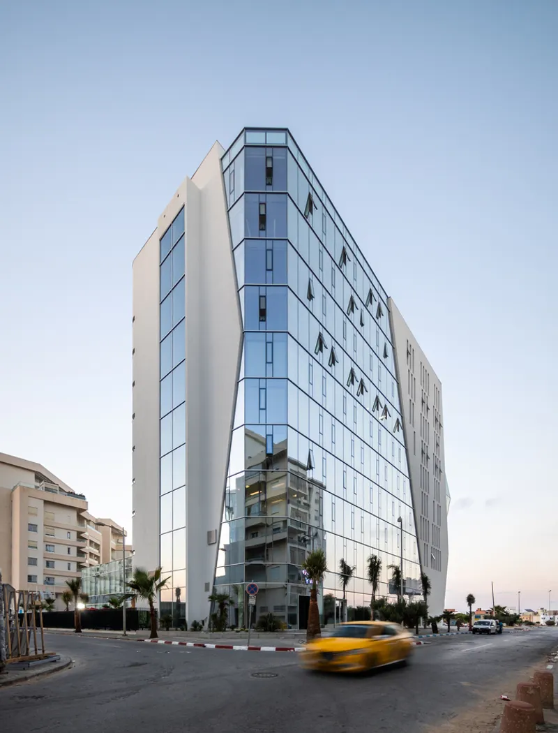 Corner elevation of glass and white panel facade at dusk with yellow taxi passing in foreground