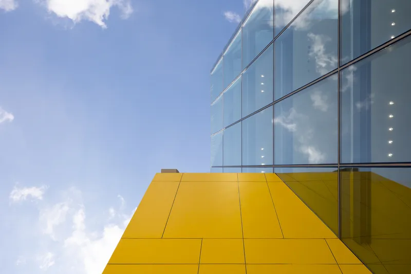 Upward view of yellow metal panel soffit meeting glass curtain wall against blue sky with clouds