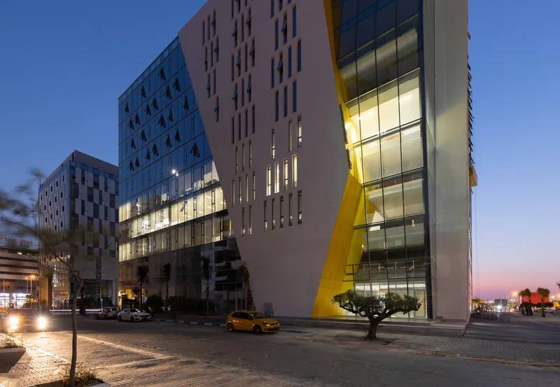 Street view of mixed-use towers with yellow diagonal brace and illuminated lobbies at sunset