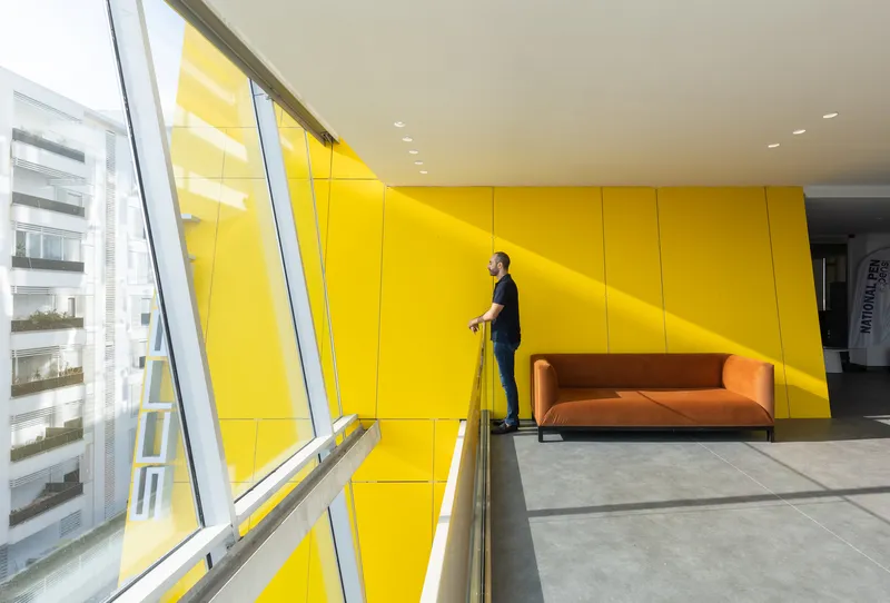 Yellow paneled wall beside the slanted glazing with a person standing near an orange sofa in sunlight