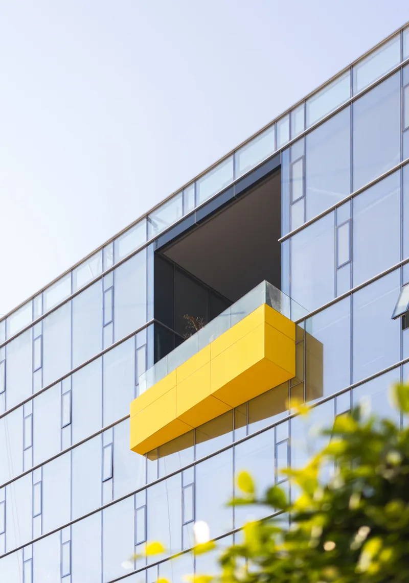 Yellow cantilevered balcony projecting from the white paneled facade with foliage visible below