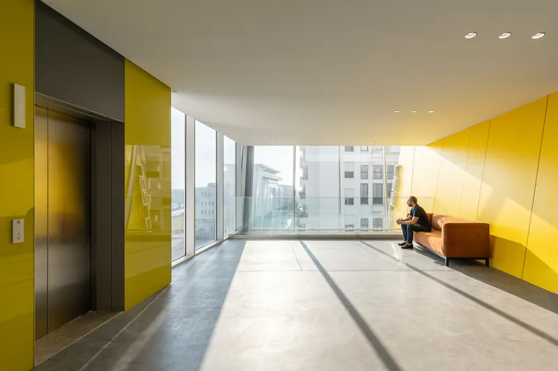 Elevator lobby with yellow wall panels and slanted floor-to-ceiling windows casting angular shadows across the floor
