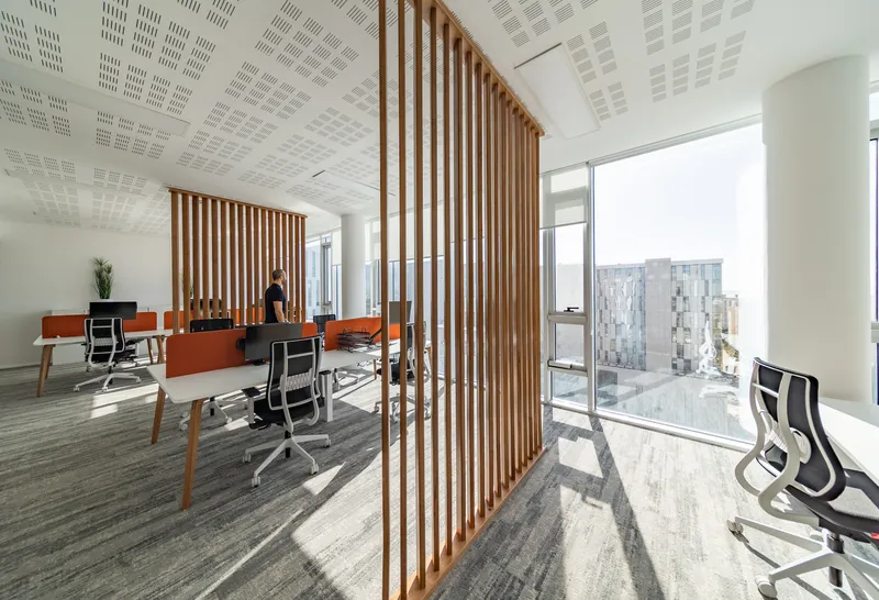 Workspace divided by vertical timber slat screens with a person standing near workstations beneath perforated acoustic ceiling
