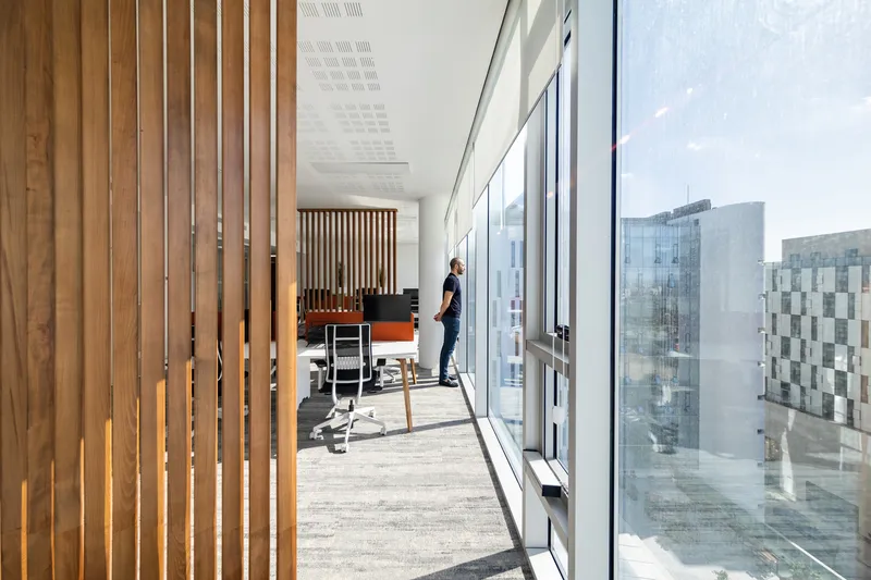 Glazed corridor with timber slat partitions and a figure near the window overlooking neighboring buildings in bright daylight
