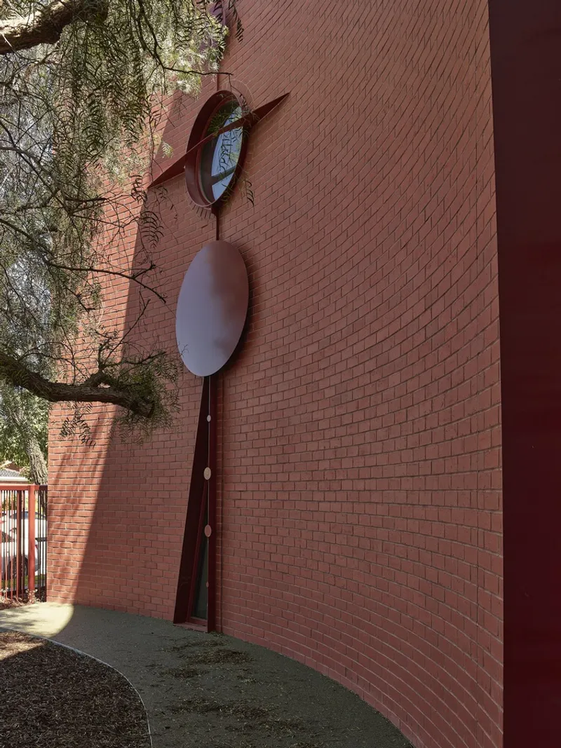 Tall brick wall with vertical metal sculpture featuring circular mirror and oval panels beneath tree branches