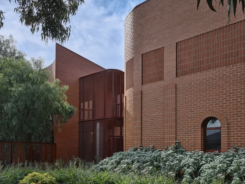 Corner detail of cylindrical brick tower with circular window beside curved metal panel volume and planted foreground
