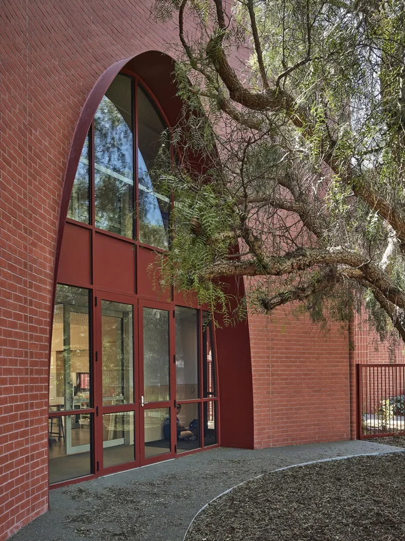 Arched entrance with glazed metal doors set into red brick facade beneath overhanging tree limbs