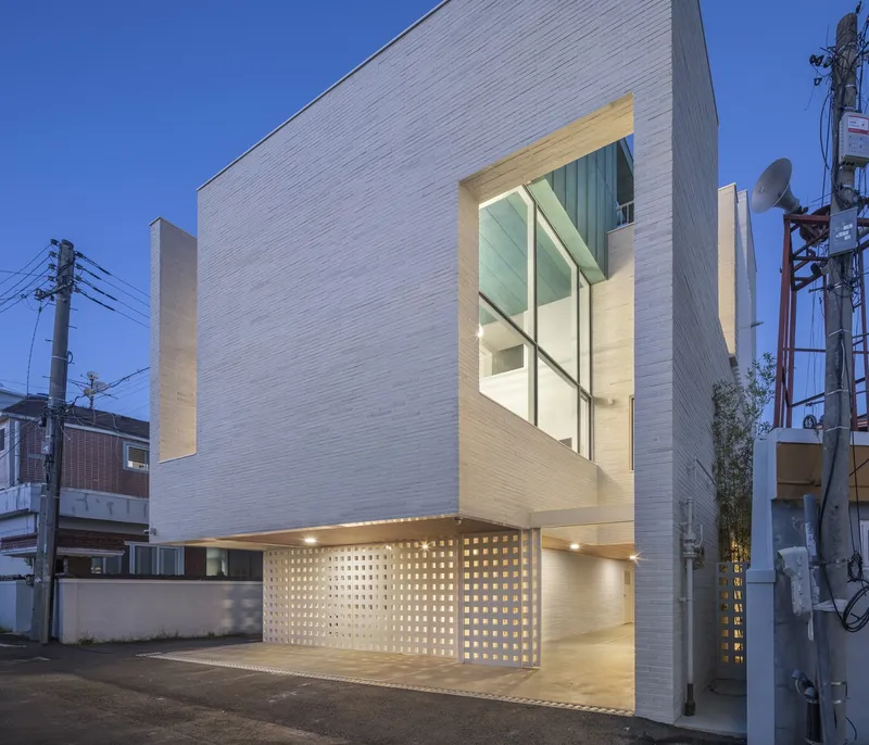 Street facade with horizontal white cladding and a perforated screen at the illuminated ground level at dusk